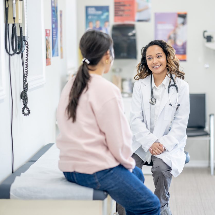 A middle aged woman of Asian decent, sits up on an exam table as she talks with her female doctor about her health concerns.